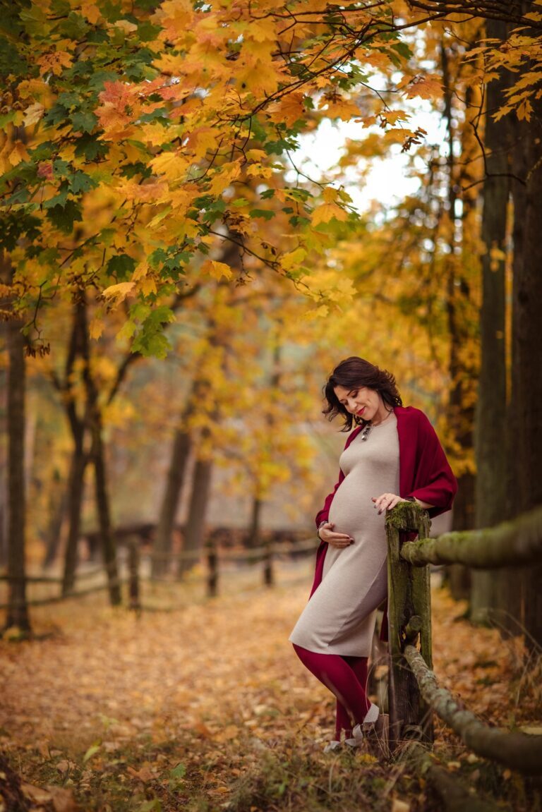 Expectant mother in a burgundy wrap leaning on a fence under golden maples