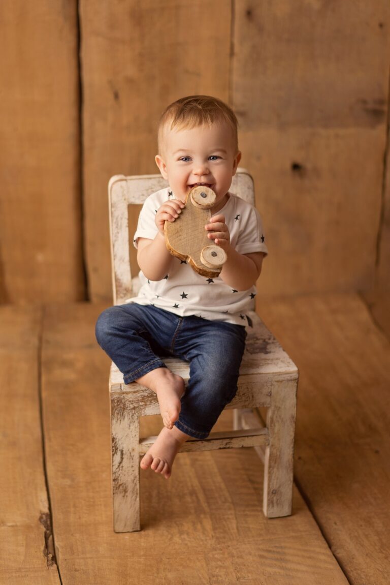 Smiling baby in jeans on a white wooden chair biting a wooden toy car