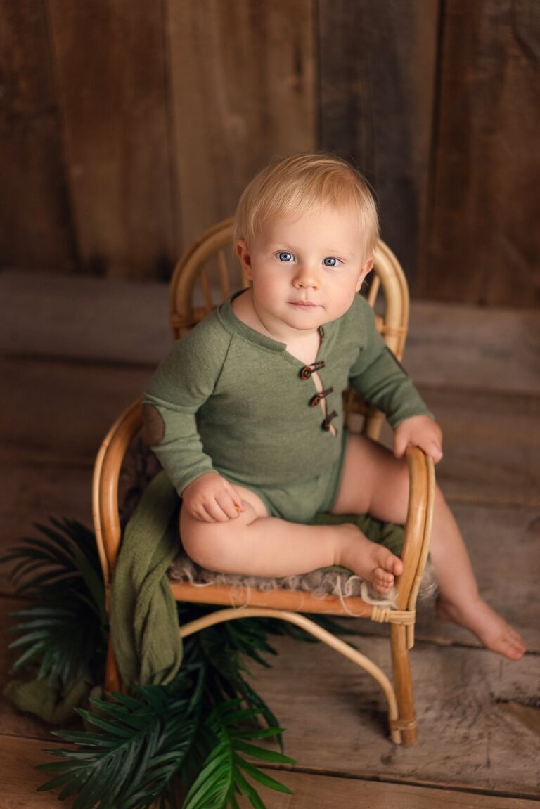 Blond baby in a green button romper on a rattan chair with palm leaves