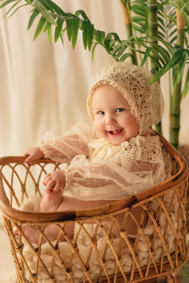 Laughing baby in a lace bonnet and dress in a rattan crib by palms