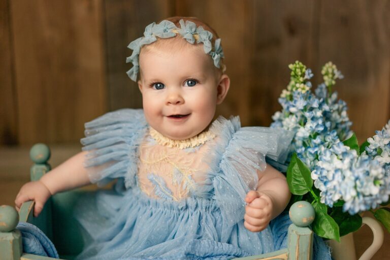 Smiling baby girl in a blue tulle dress and floral headband beside hydrangeas