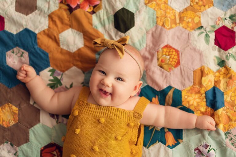 Laughing baby girl in a mustard knit romper lying on a hexagon patchwork quilt