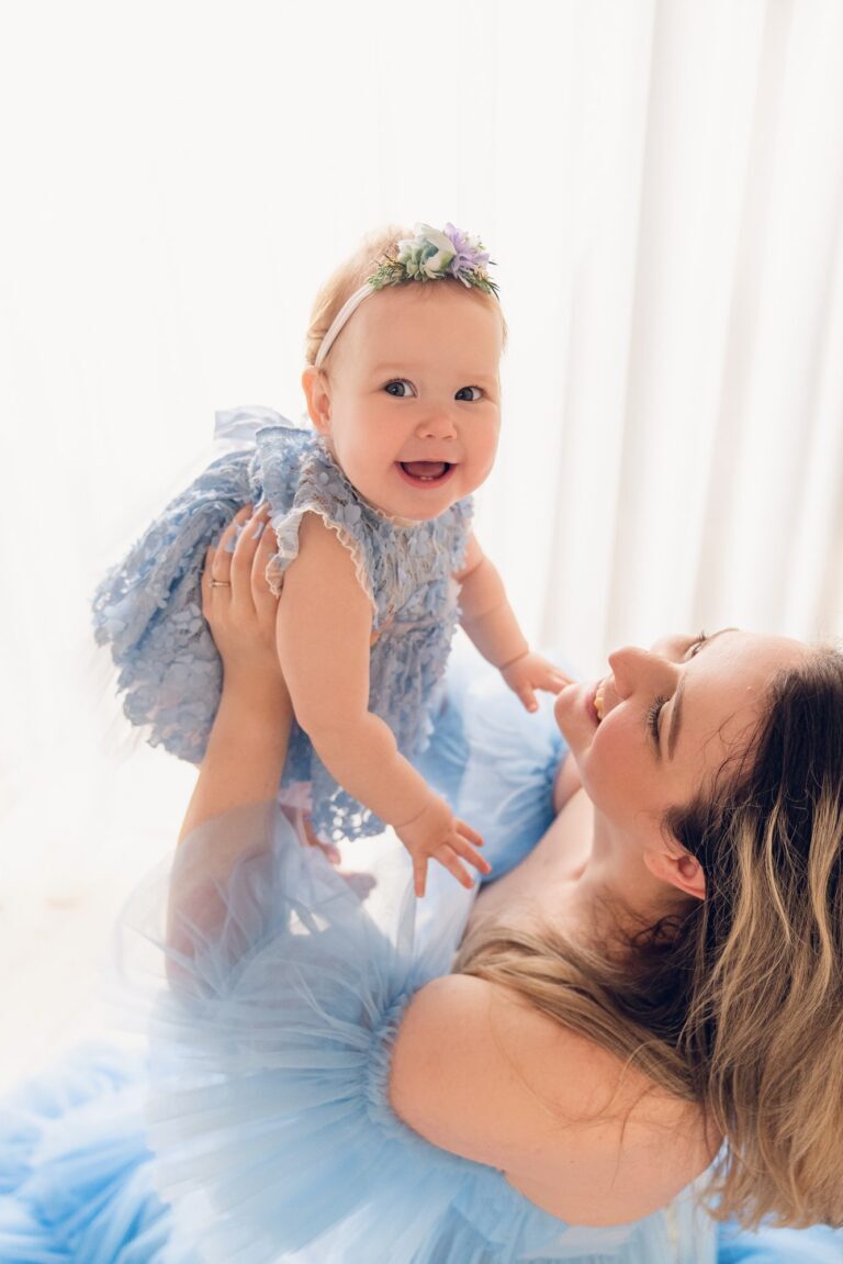 Mother in a blue tulle gown lifting her laughing baby in a lace dress