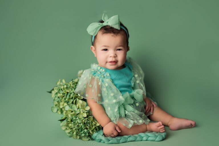 Baby girl in a mint polka-dot tulle dress and bow beside green hydrangeas