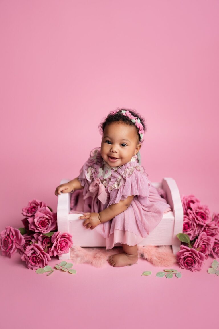 Smiling baby girl in mauve ruffles perched on a pink wooden bed among roses
