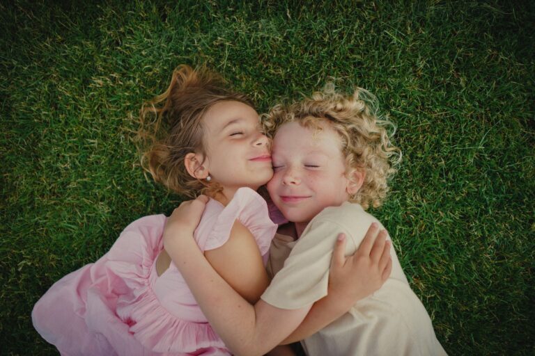 Two smiling siblings hugging with eyes closed while lying on green grass