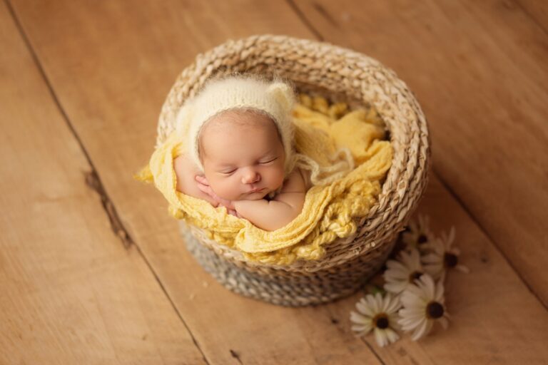 Newborn in a cream bear bonnet asleep in a woven basket beside daisies