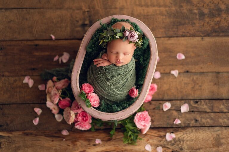 Newborn girl in a sage wrap and floral crown asleep in a pink bowl