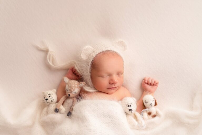 Sleeping newborn in a white bear-ear bonnet beside four little knit animal toys