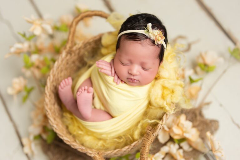Newborn girl in a yellow wrap asleep in a woven basket among cream flowers