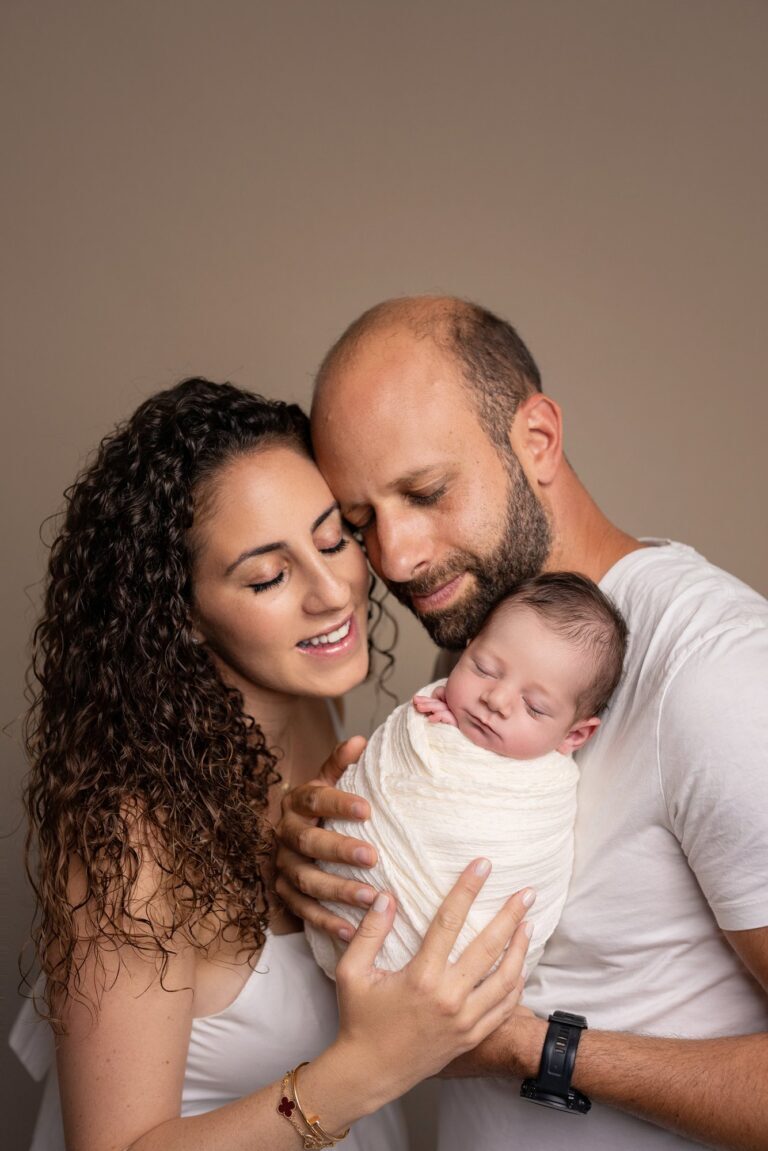 Parents cheek to cheek cradling their sleeping newborn in a cream wrap