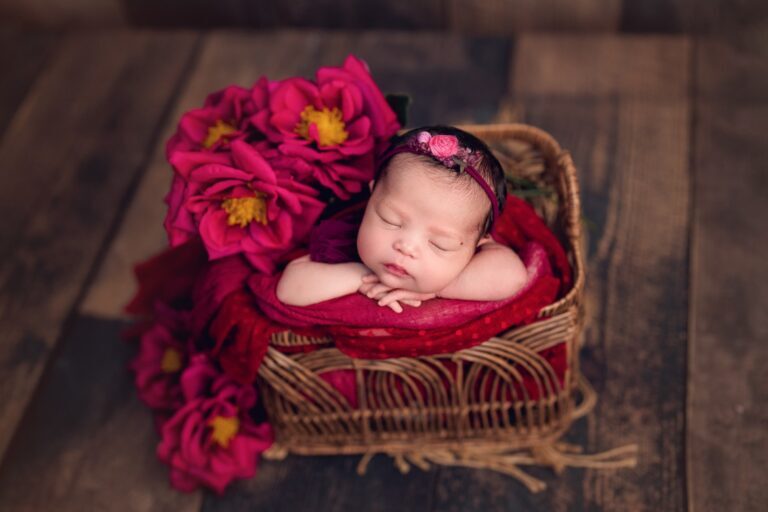 Newborn girl in a berry wrap asleep in a wicker basket beside magenta flowers
