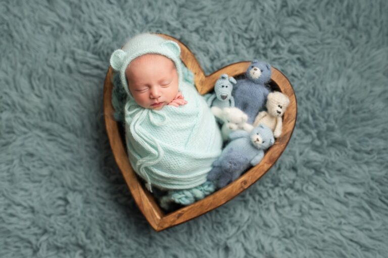 Newborn in a mint bear-ear bonnet asleep in a heart bowl of blue teddies
