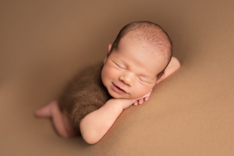 Smiling sleeping newborn in a brown knit romper with chin on hands