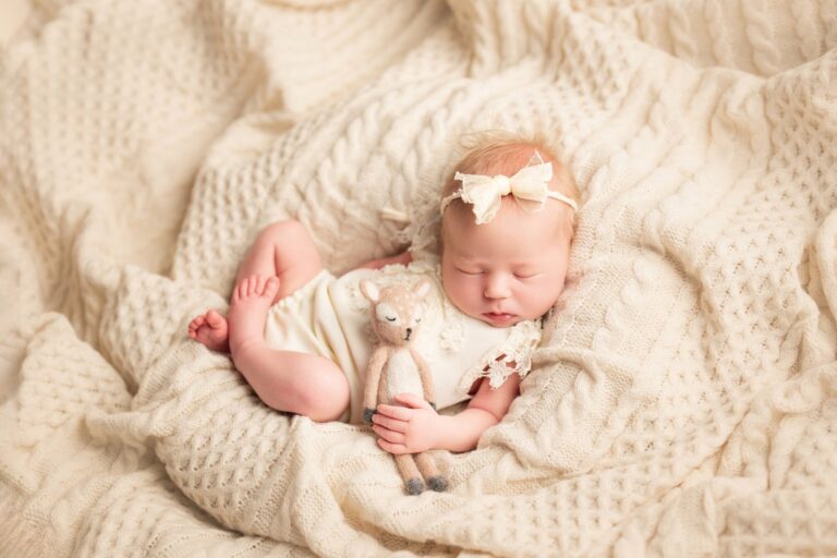 Newborn girl in cream with a bow, holding a toy fawn on cable knit