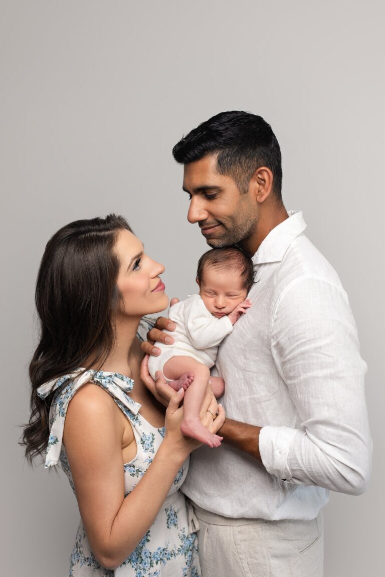 Parents gazing at each other while holding their newborn in a white knit romper