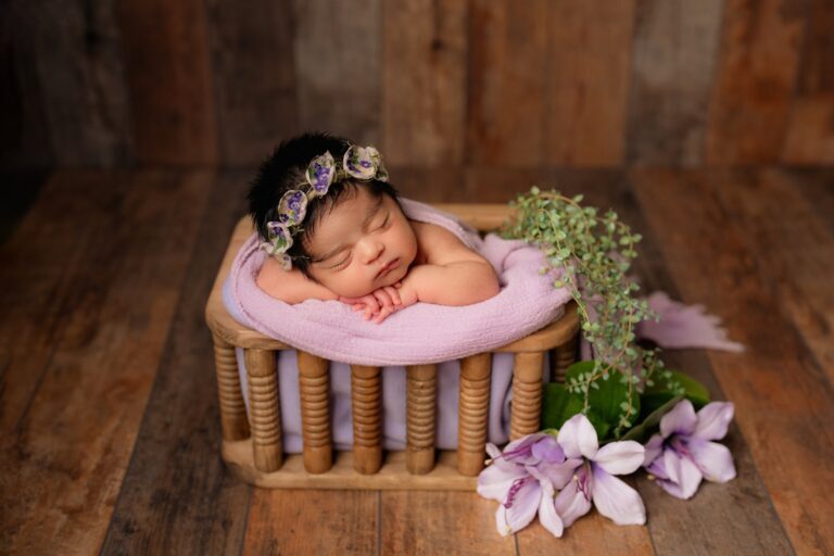 Newborn girl in a purple floral crown asleep on a spindle bed with lilies