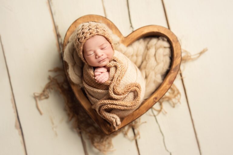 Newborn in a caramel knit bonnet and wrap asleep in a wooden heart bowl