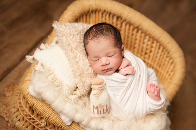 Newborn swaddled in white asleep in a wicker chair beside a felt milk bottle