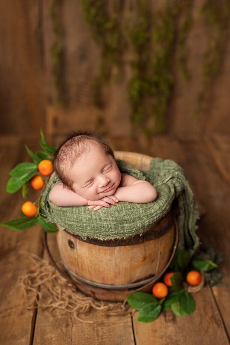 Smiling newborn asleep with chin on hands in a wooden bucket beside little oranges