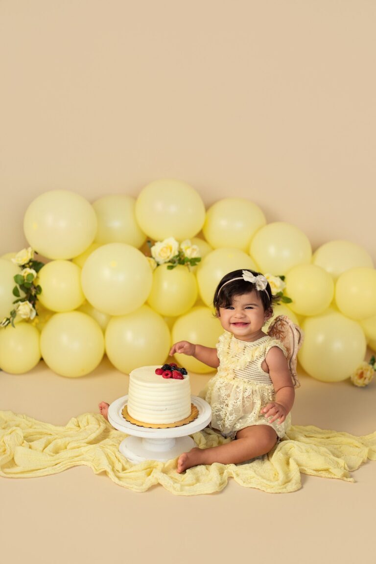 Smiling baby girl in yellow lace beside a berry cake and yellow balloons