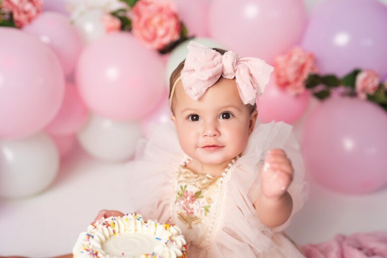 Baby girl with a big pink bow sitting behind a sprinkle-trimmed cake