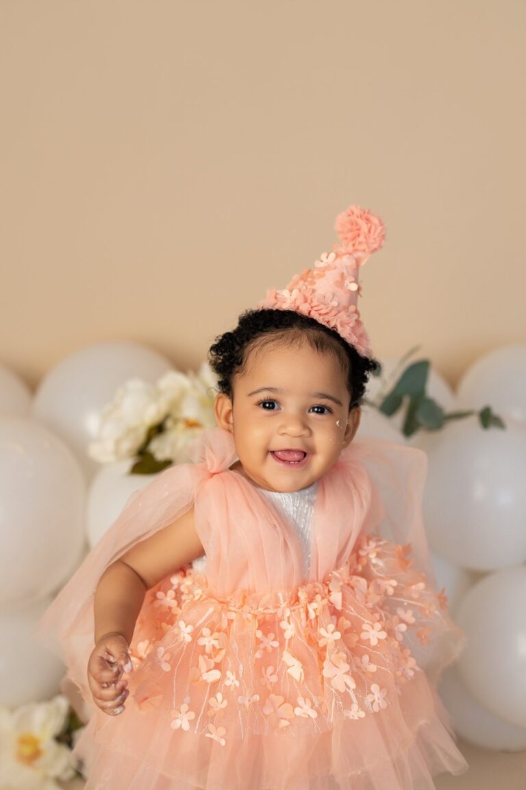 Smiling girl in a peach floral tulle dress and party hat by white balloons