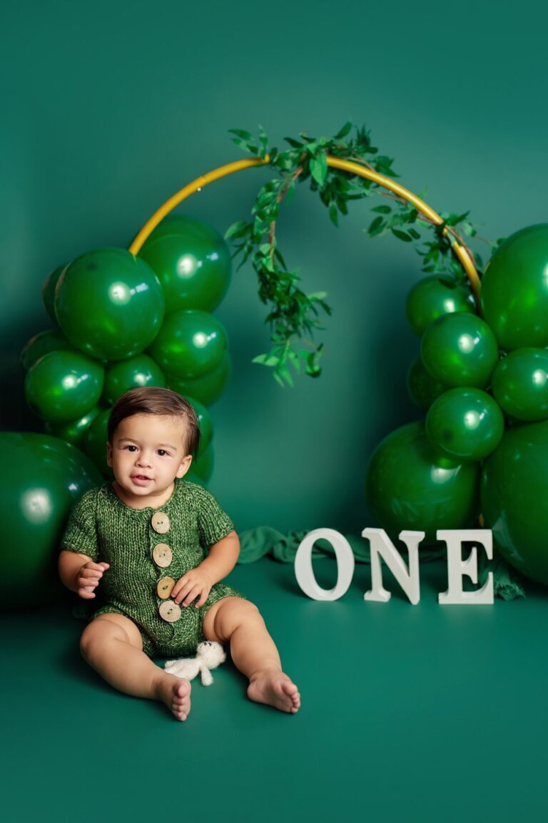 Baby in a green knit romper under an arch of green balloons