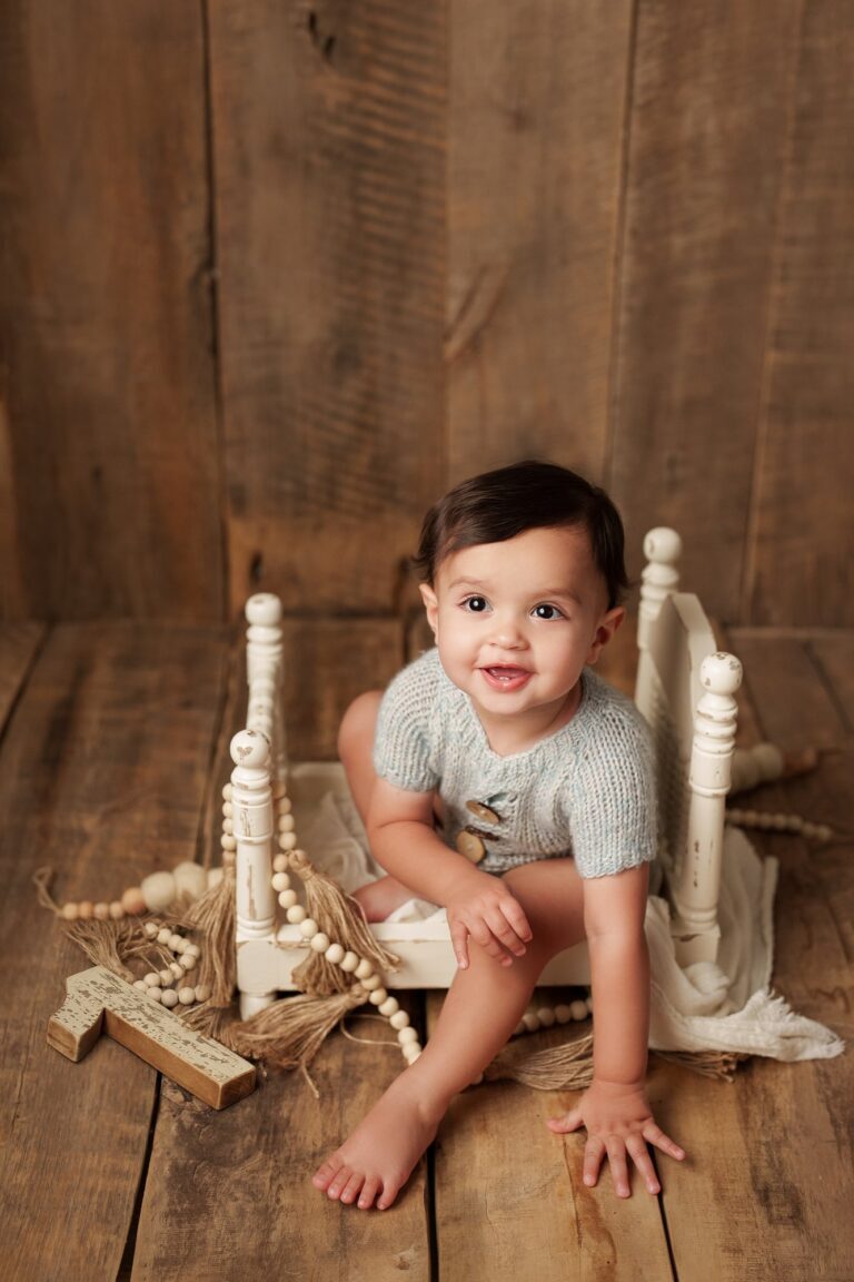 Smiling baby in a blue knit romper sitting on a white spindle bed frame