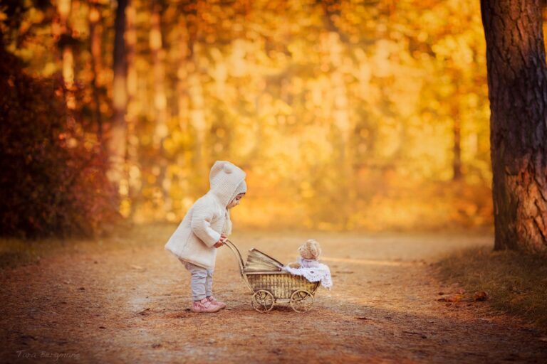 Toddler in a white bear-ear hoodie pushing a wicker pram on an autumn path