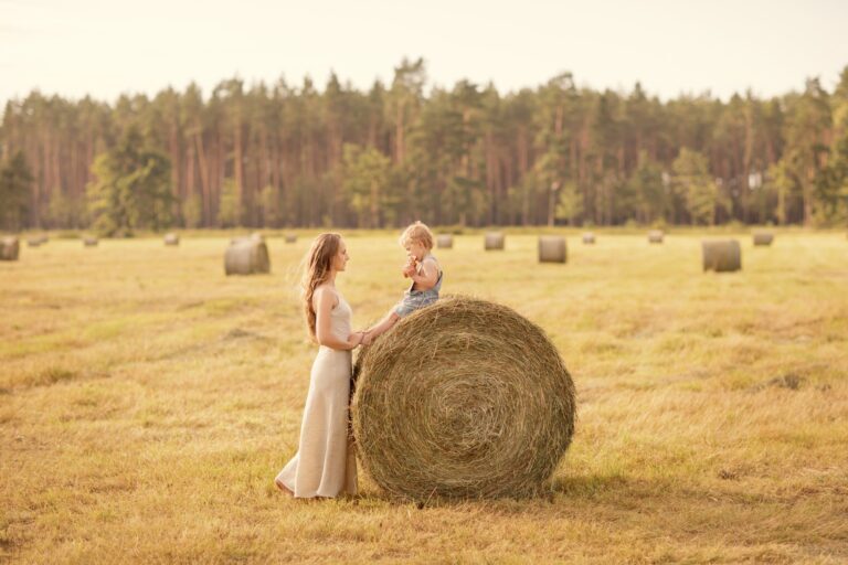 Mother holding hands with her toddler atop a round hay bale in a field