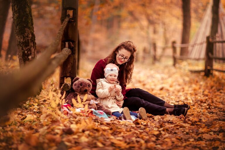 Mother and toddler sitting on autumn leaves beside a brown teddy bear