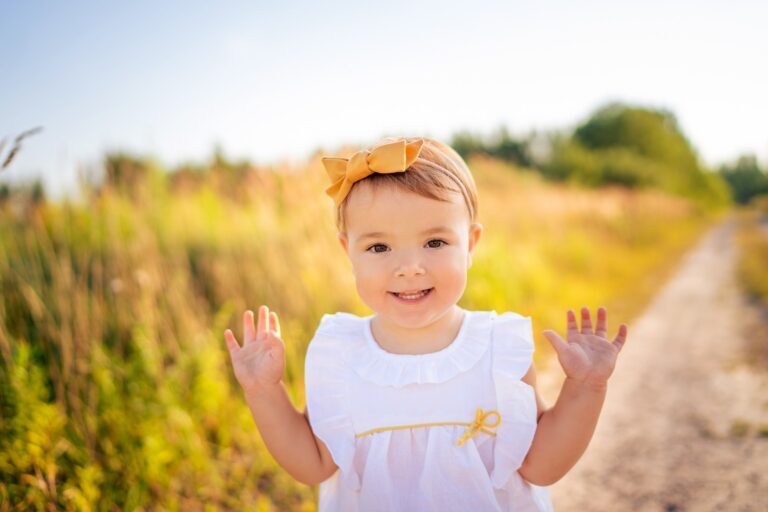 Smiling toddler girl with a mustard bow raising both hands on a country road