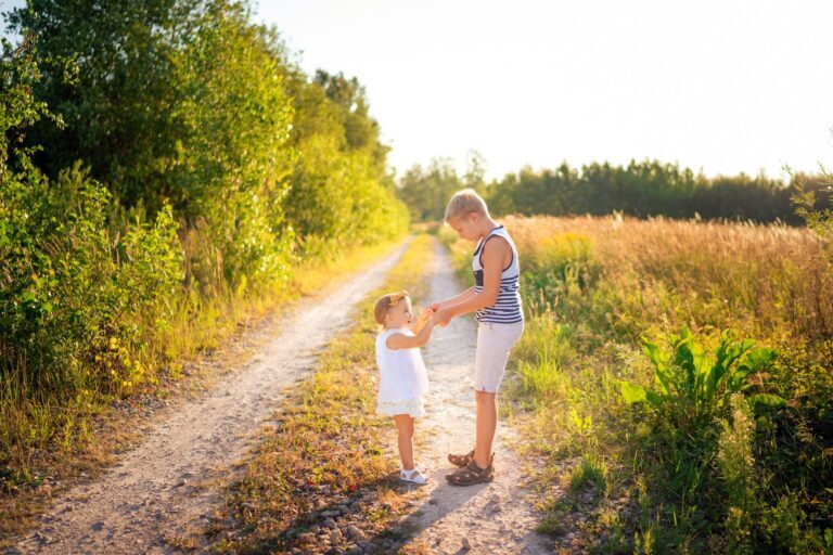 Siblings holding hands on a sunlit dirt road beside a golden field