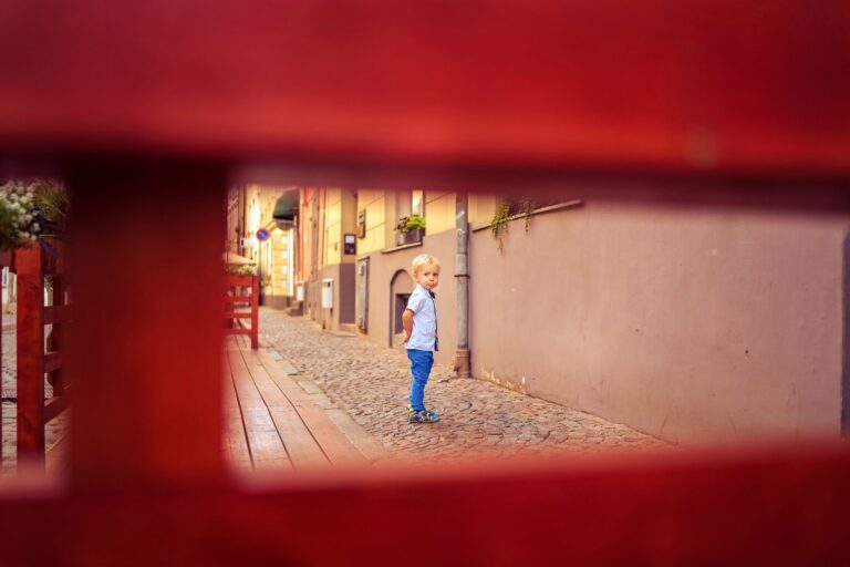 Toddler in a blue shirt on a cobblestone lane framed by a red fence