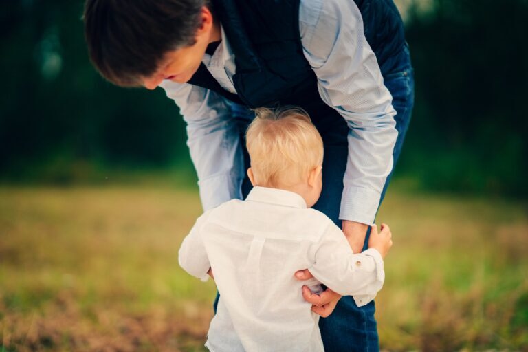Toddler boy in a white linen shirt hugging his father's legs in a field