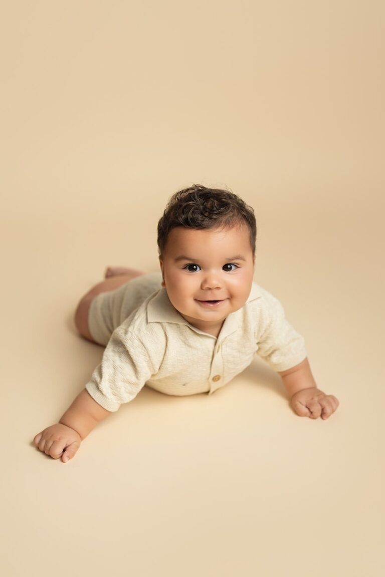 Smiling baby in a cream knit polo romper propped up on a beige backdrop