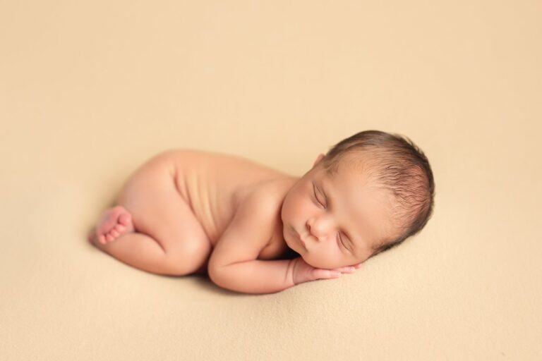 Newborn asleep on one side, hand under cheek, on a cream backdrop