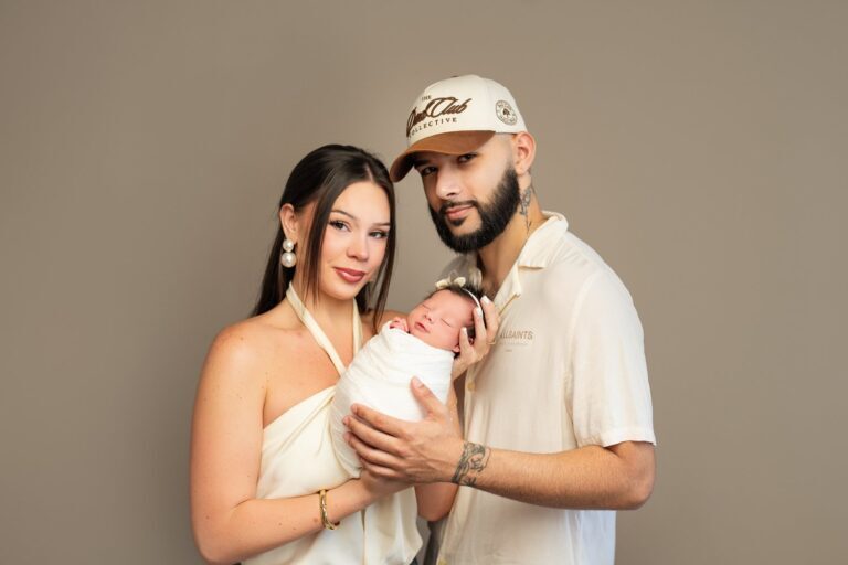 Parents in cream holding their swaddled newborn girl against a taupe backdrop