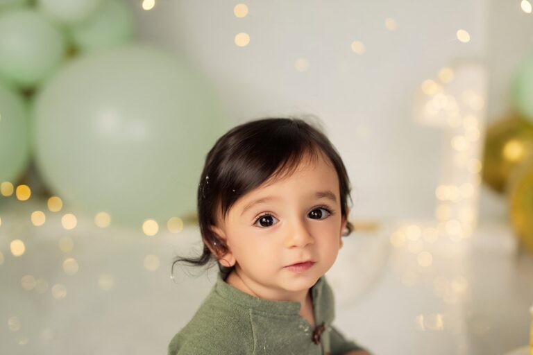 Dark-haired baby in a green knit top among mint balloons and twinkle lights
