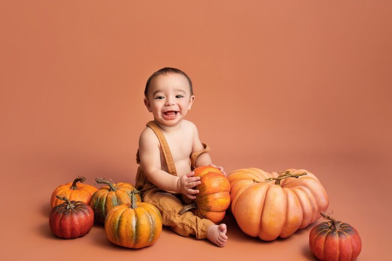 Laughing baby in tan overalls holding a pumpkin on a terracotta backdrop