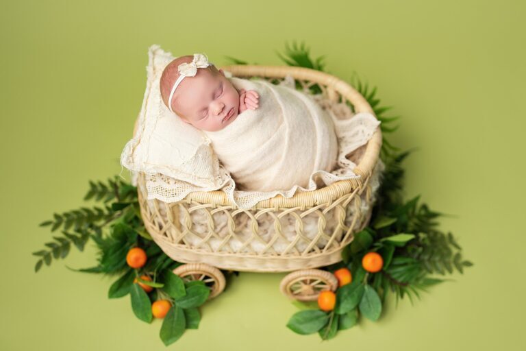 Newborn girl in a bow headband, swaddled in cream, in a rattan pram with tangerines