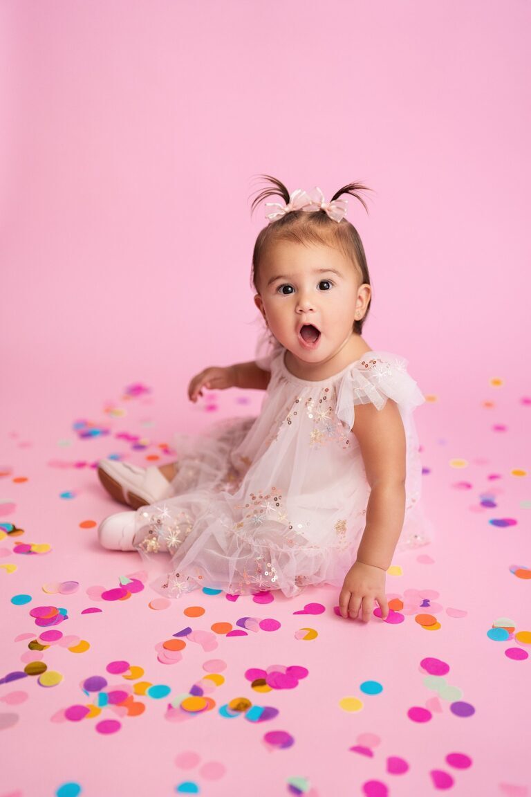 Baby girl with pigtails in a white sequin tulle dress sitting in colorful confetti