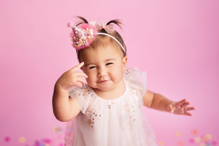 Squinting baby girl in a pink party hat pointing a finger on a pink backdrop