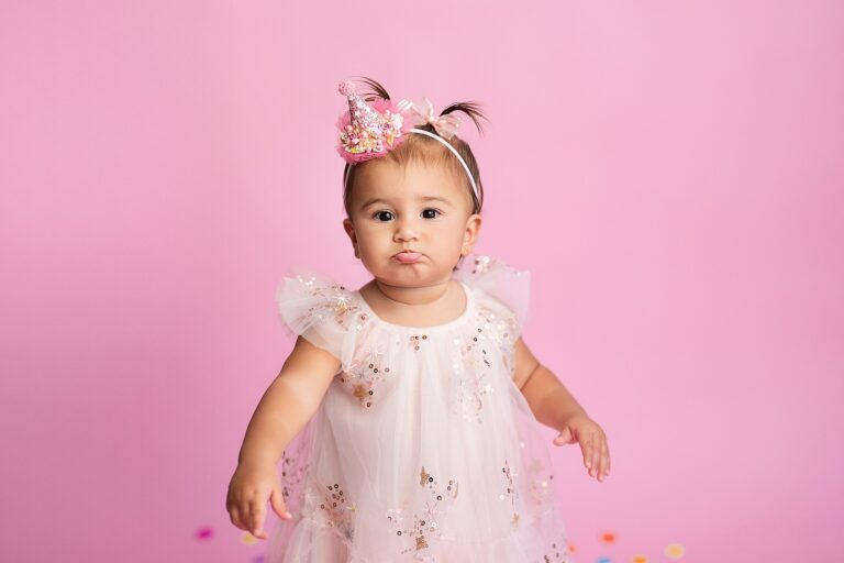 Pouting baby girl standing in a pink party hat and white sequin tulle dress
