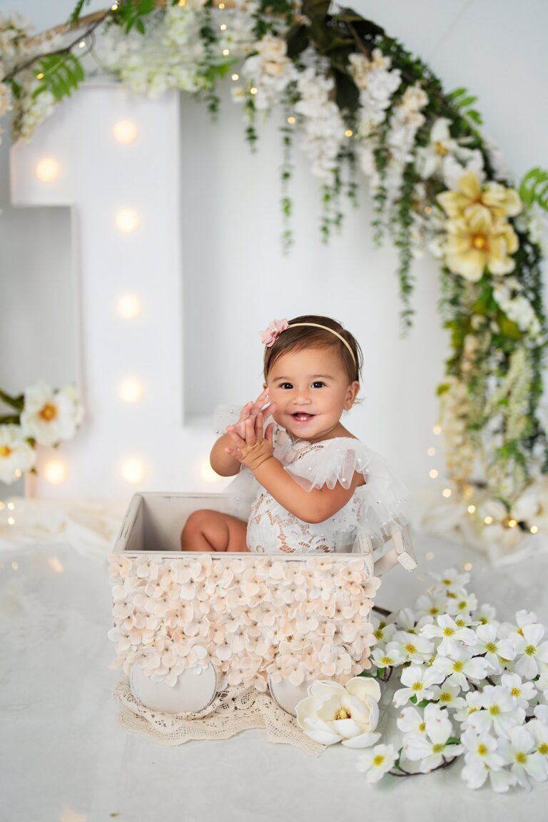 Baby girl in white lace clapping in a flower-covered wagon by a lighted number one