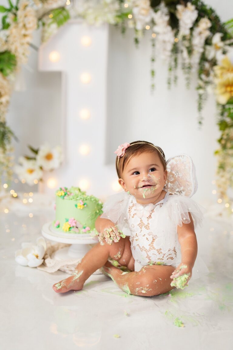 Baby girl in white lace and fairy wings, covered in green frosting, by her cake